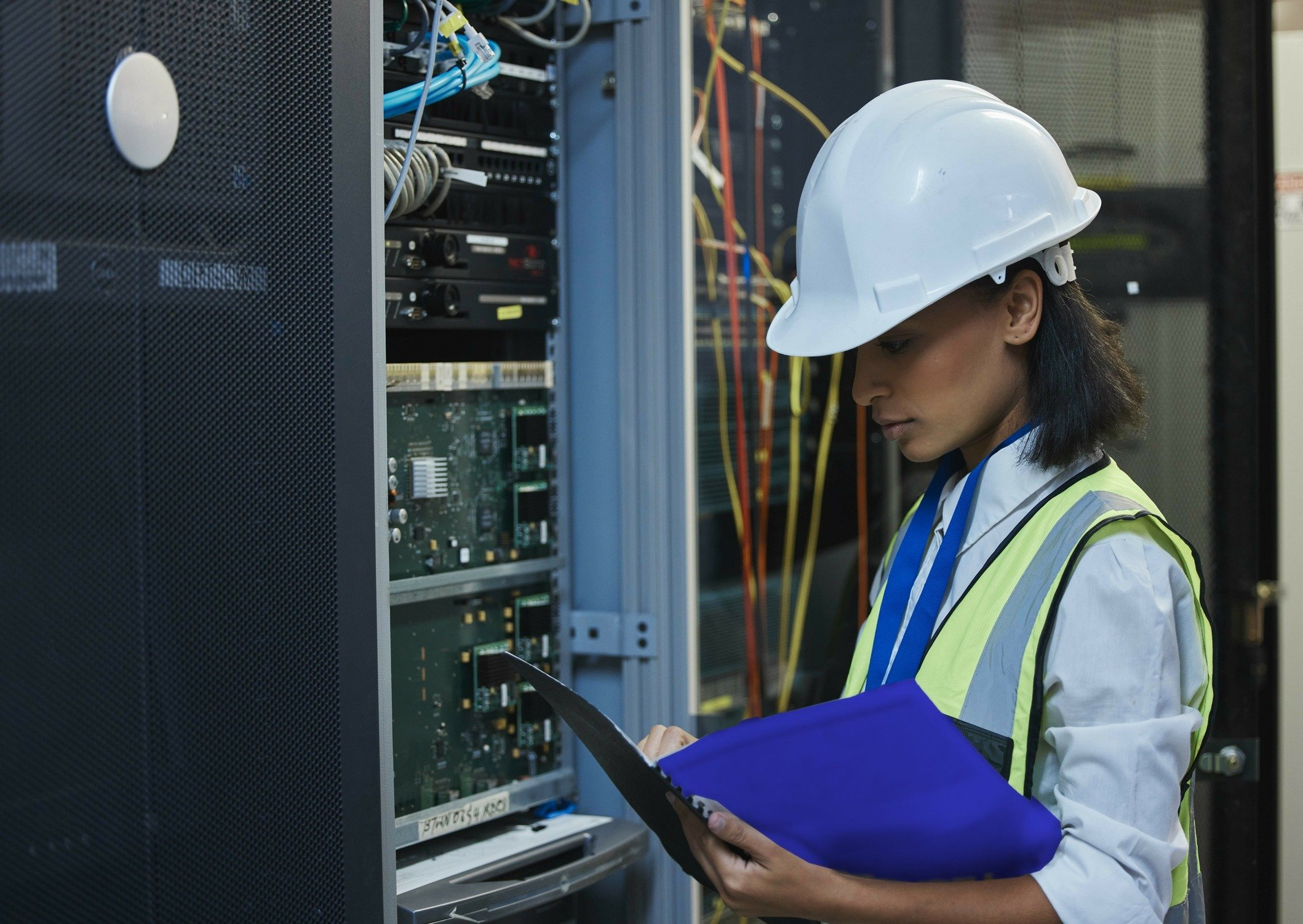 A technician in a hard hat reads a printed procedure beside a server rack